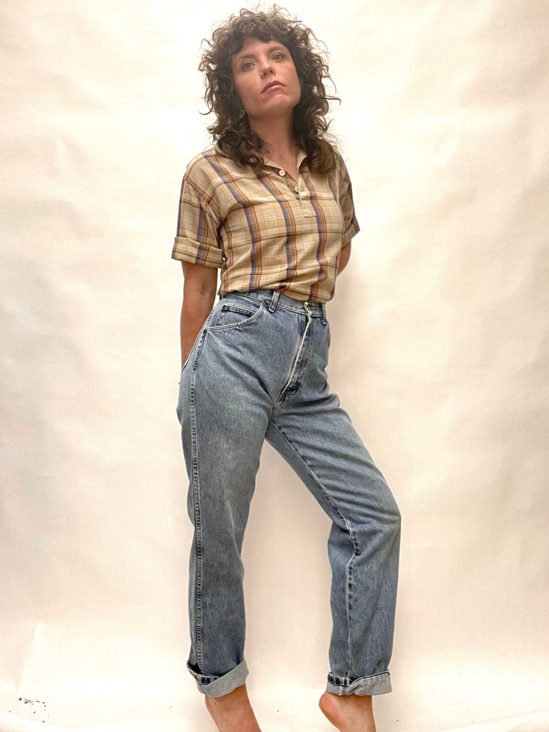 Op de afbeelding: Een vrouw draagt een lichtbruin en blauw geruit overhemd met korte mouwen en een lichtblauwe jeans met hoge taille.