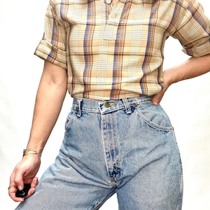 Op de afbeelding: Een beige en blauw geruit overhemd met korte mouwen, een kraag en een enkele knoopsluiting. Het overhemd wordt gedragen met lichtblauwe jeans.