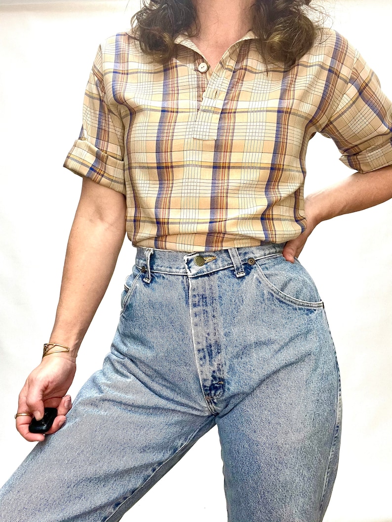 Op de afbeelding: Een beige en blauw geruit overhemd met korte mouwen, een kraag en een enkele knoopsluiting. Het overhemd wordt gedragen met lichtblauwe jeans.