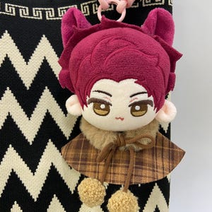 May include: A plush doll keychain featuring a character with short, deep magenta hair, pointed ears, and embroidered brown eyes. The doll wears a brown plaid cape with a tan faux fur collar and two small tan pom-poms hanging from a brown drawstring.