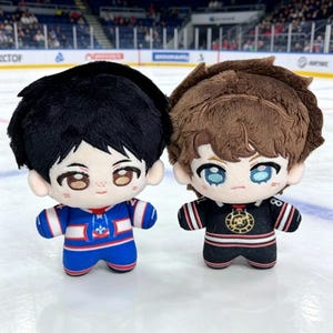 May include: Two plush dolls with anime-style features. One doll wears a blue and white hockey jersey, the other a black and red jersey. Both dolls have large eyes and fluffy hair, set against an ice rink background.
