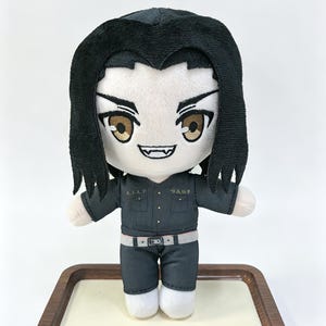 May include: A plush doll featuring a character with black hair, brown eyes, and visible fangs. The doll wears a dark gray button-up uniform with a light gray belt and white shoes. The character has a wide, smiling expression with sharp teeth.