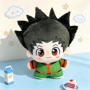 May include: A plush doll featuring a character with spiky dark green hair, large orange eyes, and a small smile. The doll wears a green jacket with orange trim and matching green pants. It has a soft texture and a rounded, chibi-style body shape.