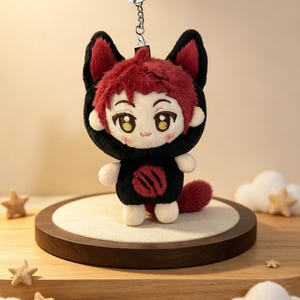 May include: A plush keychain featuring a character with short red hair and large golden eyes, wearing a black hooded outfit with red animal ears and a red circular emblem on the chest. The figure has a small red tail and a metal chain attachment.