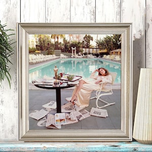 May include: Framed print of a woman lounging by a pool, surrounded by newspapers. The image is in a silver frame, set on a blue and white surface. A vase and a black and white polka-dot cup are also on the surface.