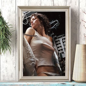 May include: Framed print featuring a woman in a white tank top and underwear. The image is set against a dark background, framed in a silver-toned frame. A vase and a decorative black and white container are on a blue wooden surface.