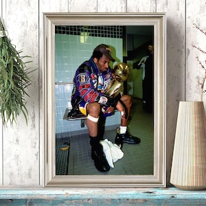 Puede incluir: Fotografía enmarcada de una persona sosteniendo un trofeo dorado. La persona lleva una chaqueta morada y amarilla y una gorra de béisbol. La fotografía está en un marco plateado y se muestra en un estante azul y blanco.