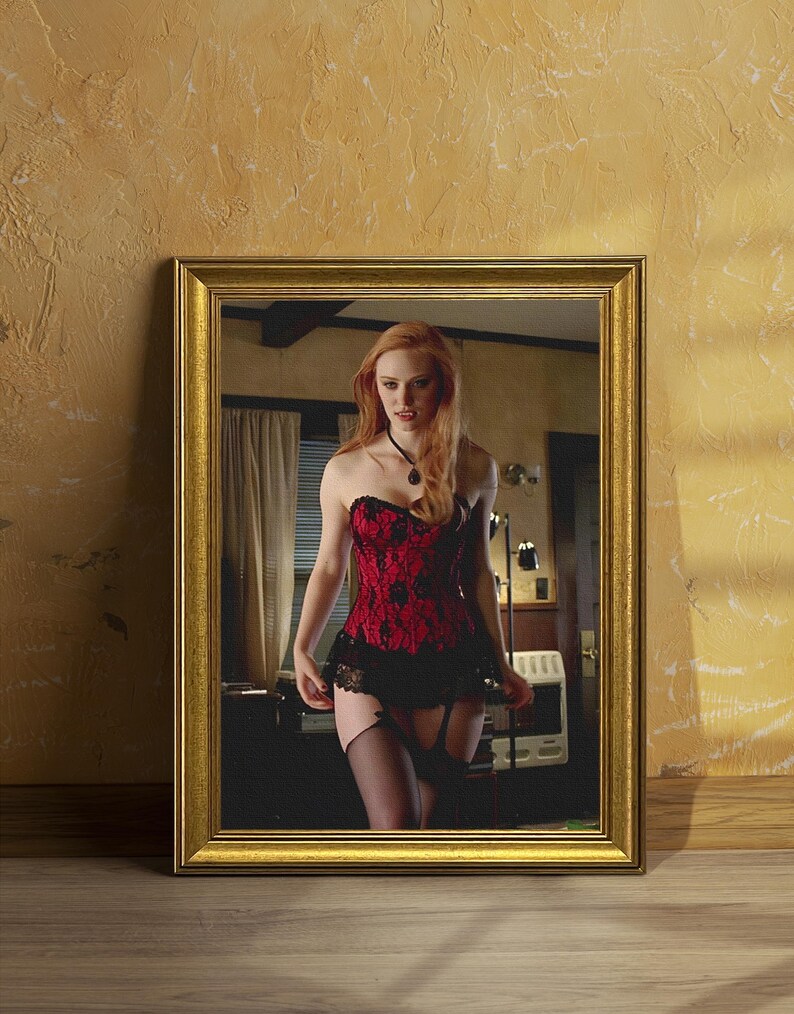 May include: A framed mirror with a gold-coloured frame. The mirror reflects a person wearing a red and black corset with black stockings. The background is a room with a desk and curtains.