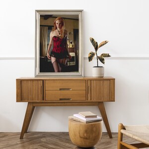 May include: A wooden cabinet in a mid-century modern style, featuring two doors, two drawers, and angled legs. A mirror above reflects a person in a red and black corset. A small wooden stool and a potted plant are also visible in the room.