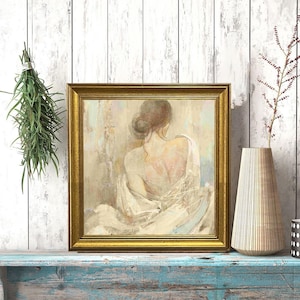 May include: A framed artwork with a gold frame shows a woman from the back, in soft, muted tones. The artwork is on a distressed blue shelf, with a vase and a black and white dotted container. Greenery hangs on the left.