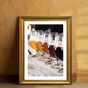 May include: A framed print featuring a beer flight with five different beers in tulip glasses. The beers range in color from light golden to dark brown, showcasing the variety of brews. The glasses are arranged on a wooden surface.