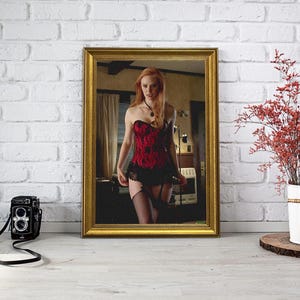 May include: A framed image of a woman in a red and black corset, stockings, and garter. A vintage black camera with a strap is in front of the frame. A white bucket with red branches sits on a wooden base.