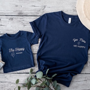 May include: Two navy blue t-shirts, one adult and one child size, with white text. The adult shirt says "You Make ME HAPPY" and the child's shirt says "The Happy MAKER". A straw hat and toy camera are also in the image.