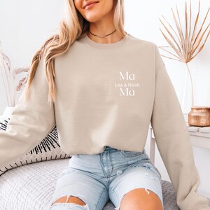 May include: Beige crewneck sweatshirt with the word "Ma" above "Lea & Noah Ma" in white lettering. The sweatshirt is a neutral color and has long sleeves. The image is of a person wearing the sweatshirt.