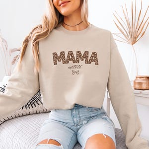 May include: Beige crewneck sweatshirt with the word "MAMA" in a brown leopard print design. Below the word "MAMA" is the text "est 2026" and a small bow graphic. The sweatshirt is a neutral color.