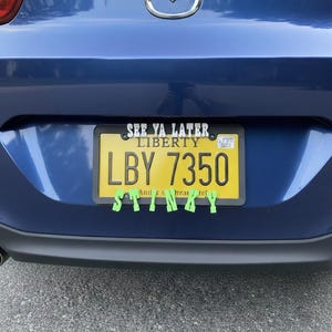 May include: A blue car's rear with a yellow license plate that reads "LBY 7350" and "SEE YA LATER LIBERTY". Green sticky notes spell "STINKY" across the bottom of the plate.
