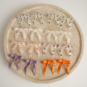 Könnte beinhalten: Eine Sammlung von ohrringförmigen Schleifen in verschiedenen Farben und Mustern. Die Ohrringe weisen Designs mit Punkten, floralen Drucken und Volltonfarben wie Lila und Orange auf. Sie werden auf einem beigen, kreisförmigen Tablett präsentiert.