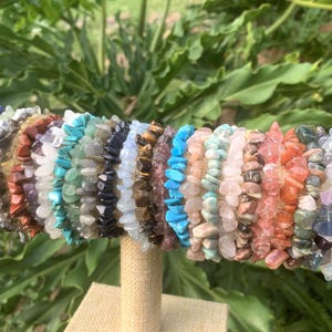 May include: A selection of gemstone chip bracelets in various colours, including blue, green, brown, and purple. The bracelets are displayed on a beige cylindrical stand against a backdrop of green foliage.