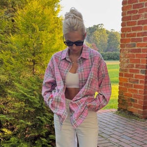 Könnte beinhalten: Ein rosa kariertes Hemd mit langen Ärmeln, kombiniert mit einer weißen Hose. Das Hemd hat ein klassisches Karomuster in Rosa und Grau. Das Outfit wird durch ein weißes Oberteil ergänzt.