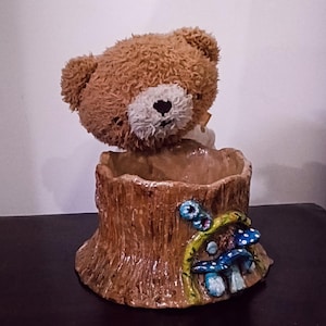 Puede incluir: Una maceta caprichosa de oso de peluche marrón. La maceta tiene forma de tocón de árbol con detalles de setas azules y blancas. La cabeza del oso está colocada encima de la maceta, añadiendo un toque juguetón a este artículo decorativo.