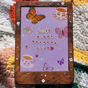 Könnte beinhalten: Eine E-Reader-Hülle mit Holzrahmen und einem lavendelfarbenen Hintergrund mit Schmetterlingen, Teetassen und dem Text "LATE NIGHT READERS CLUB". Die Hülle ist mit kleinen weißen Blumen und Edelsteinen verziert.