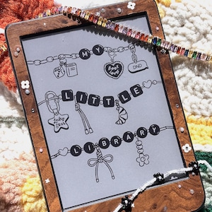 Peut inclure: Liseuse à cadre en bois avec les mots "MY LITTLE LIBRARY" et des illustrations de breloques sur l'écran. Une guirlande de perles colorées et une guirlande de perles blanches avec des accents de fleurs noires sont drapées autour de la liseuse, posée sur une couverture crochetée colorée.