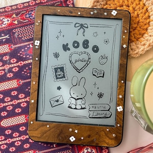 以下が含まれることがあります： 木製のフレームに小さな白い花が飾られた電子書籍リーダーが、模様入りのラグの上に置かれています。「girlie」という言葉とウサギの落書きが画面に表示されています。 かぎ針編みのコースターとキャンドルも表示されています。