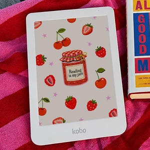 Peut inclure: Un lecteur Kobo blanc avec un motif de fraises et de cerises, et un livre de poche intitulé "Good Material" de Dolly Alderton, posé sur une serviette à rayures rouges et roses. Le lecteur affiche le texte "Reading is my jam".
