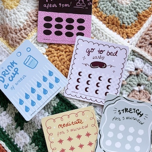 May include: A collection of reward cards with various prompts and illustrations. The cards include phrases like "Drink a cup of water," "No phone after 9 pm," "Go to bed early," "Meditate for 5 minutes," and "Stretch for 5 minutes." The cards are on a colorful crocheted blanket.