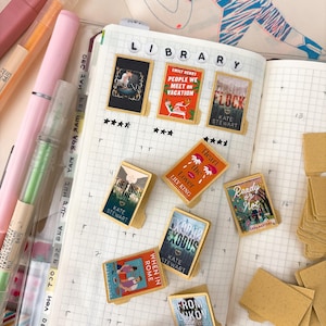 May include: A journal spread with book-shaped page markers. The markers feature book covers with titles like "People We Meet on Vacation" and "Exodus." Pens and highlighters in pink, orange, and green are visible.
