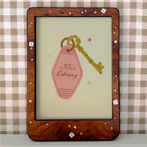 Op de afbeelding: Een rechthoekige houten fotolijst met een decoratieve rand. De lijst bevat een afbeelding van een gouden sleutel en een roze label met de tekst "MY LITTLE LIBRARY". De afbeelding heeft een crèmekleurige achtergrond met roze sterren.