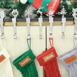 Puede incluir: Una exhibición de calcetines navideños colgados de ganchos de metal plateado. Cada calcetín es de un color diferente: blanco, verde, rojo y gris. Los calcetines tienen etiquetas con nombres personalizados. Los ganchos están decorados con adornos festivos.