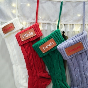 Op de afbeelding: Vier kabelgebreide kerstsokken in wit, rood, groen en lavendel. Elke sok heeft een bruin leren label met een naam geborduurd in een contrasterende kleur: Victoria, Olivia, Isabella en Charlotte. De sokken hangen aan een witte stof.