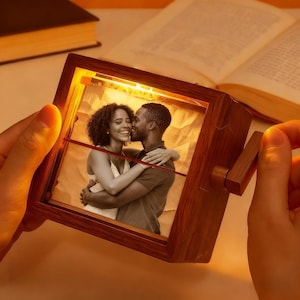 May include: A wooden photo frame with a crank, showing a black and white image of a couple embracing. The frame has a warm, glowing light. The photo is of a smiling woman and a man kissing her cheek. The frame is held by a person.