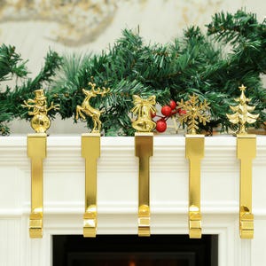 Può includere: Set di cinque portacandele natalizi color oro con disegni festivi: un pupazzo di neve, una renna, una campana, un fiocco di neve e un albero di Natale. I portacandele sono esposti su un caminetto bianco con una ghirlanda verde.