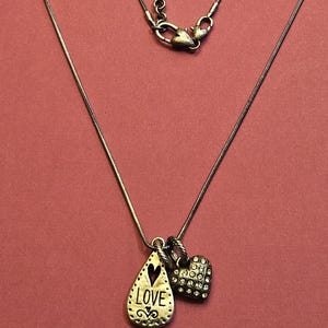 Puede incluir: Collar plateado con dos colgantes: un colgante en forma de l&aacute;grima con la palabra "LOVE" y un charm en forma de coraz&oacute;n. La cadena es delicada y tiene un cierre de mosquet&oacute;n. El fondo es de color rosa liso.
