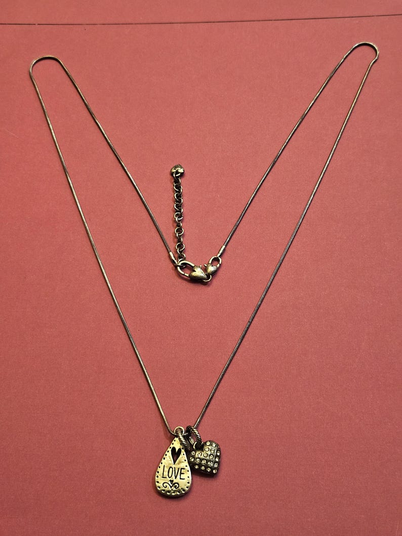 Puede incluir: Collar plateado con dos colgantes: un colgante en forma de l&aacute;grima con la palabra "LOVE" y un charm en forma de coraz&oacute;n. La cadena es delicada y tiene un cierre de mosquet&oacute;n. El fondo es de color rosa liso.