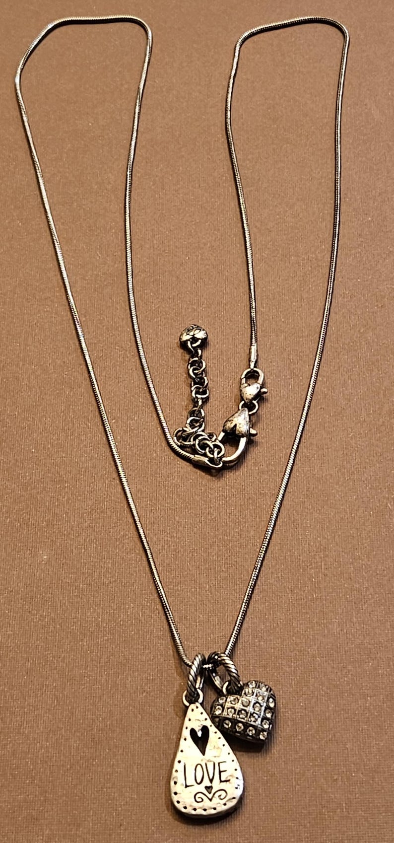 Puede incluir: Collar plateado con dos colgantes. Uno tiene forma de l&aacute;grima con la palabra "LOVE" y un coraz&oacute;n. El otro es un coraz&oacute;n cubierto de peque&ntilde;as piedras. El collar tiene una cadena fina.