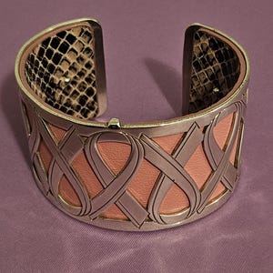 May include: Silver-tone cuff bracelet with a pink leather center and a decorative ribbon design. The interior features a snakeskin pattern. The bracelet is open at the top and bottom.