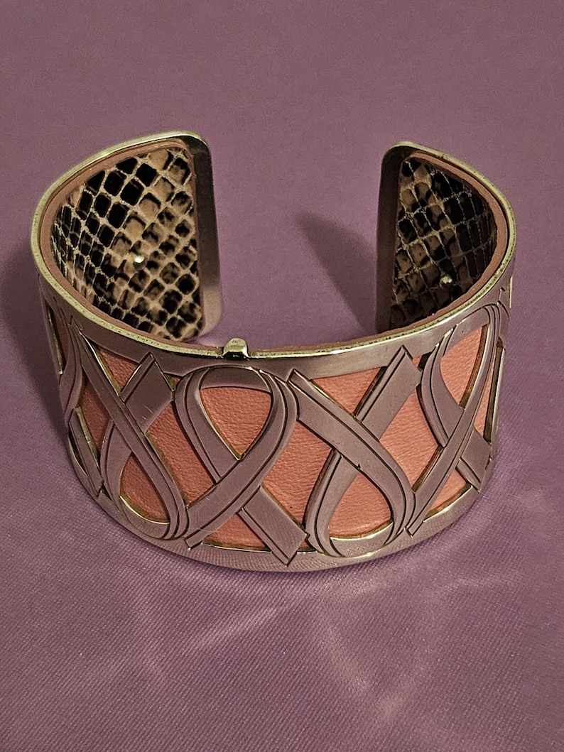 May include: Silver-tone cuff bracelet with a pink leather center and a decorative ribbon design. The interior features a snakeskin pattern. The bracelet is open at the top and bottom.