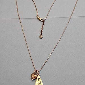 Puede incluir: Un delicado collar con una fina cadena de color bronce. Cuenta con dos colgantes: un charm en forma de coraz&oacute;n cubierto de peque&ntilde;os cristales y un colgante en forma de l&aacute;grima con la palabra "LOVE" y un coraz&oacute;n recortado. El collar se muestra sobre una superficie azul.