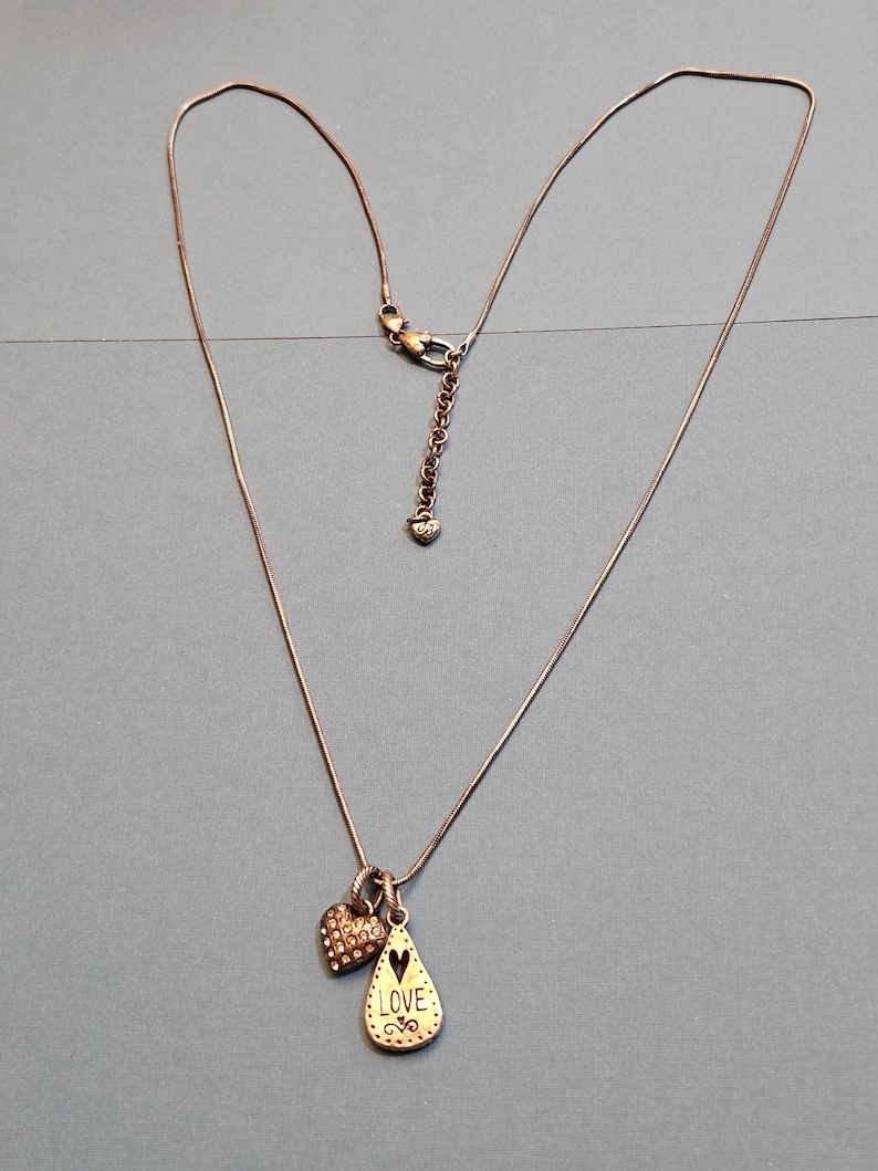 Puede incluir: Un delicado collar con una fina cadena de color bronce. Cuenta con dos colgantes: un charm en forma de coraz&oacute;n cubierto de peque&ntilde;os cristales y un colgante en forma de l&aacute;grima con la palabra "LOVE" y un coraz&oacute;n recortado. El collar se muestra sobre una superficie azul.