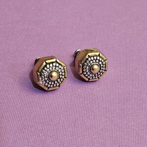 May include: A pair of octagonal stud earrings. The earrings have a gold-colored frame with a silver-colored center. The center of each earring has a small gold-colored dot. The earrings are on a purple background.