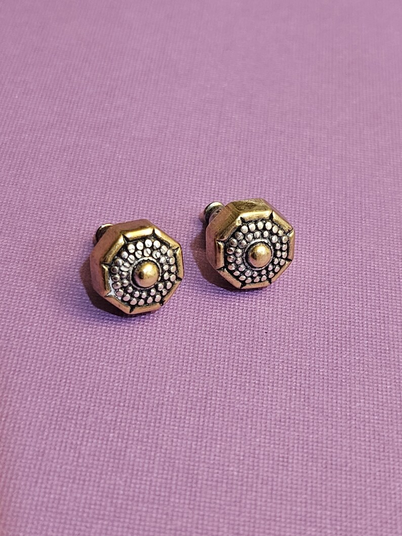 May include: A pair of octagonal stud earrings. The earrings have a gold-colored frame with a silver-colored center. The center of each earring has a small gold-colored dot. The earrings are on a purple background.