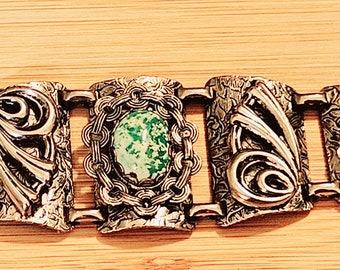 Op de afbeelding: Een zilveren armband met een groene steen in een decoratief frame. De armband heeft een gedetailleerd bloemenontwerp en een sluiting.