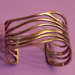 Op de afbeelding: Een goudkleurige manchetarmband bestaande uit vijf parallelle, golvende metalen banden die om de pols wikkelen. Het open ontwerp heeft een gepolijst, reflecterend oppervlak met gladde, gebogen randen over de gehele structuur.
