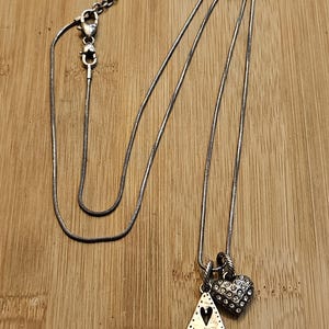 Puede incluir: Collar plateado con dos colgantes. Uno es en forma de l&aacute;grima con la palabra "LOVE" y un coraz&oacute;n. El otro es en forma de coraz&oacute;n y est&aacute; cubierto de peque&ntilde;as piedras transparentes. El collar est&aacute; sobre una superficie de madera.