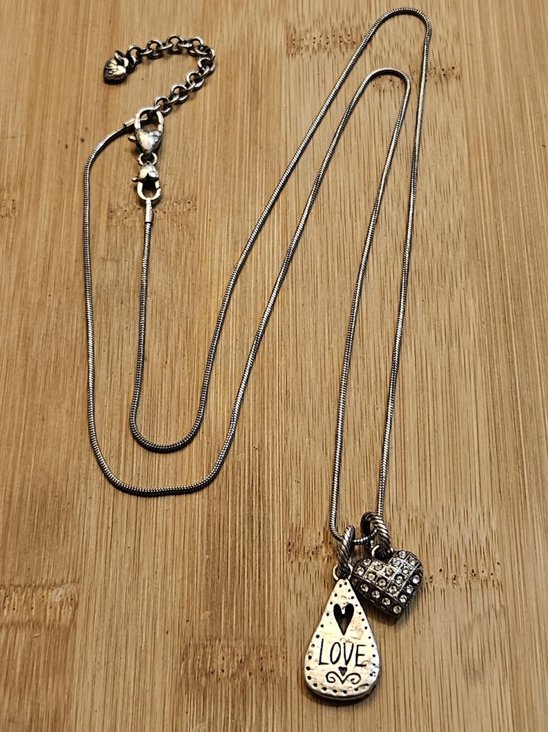 Puede incluir: Collar plateado con dos colgantes. Uno es en forma de l&aacute;grima con la palabra "LOVE" y un coraz&oacute;n. El otro es en forma de coraz&oacute;n y est&aacute; cubierto de peque&ntilde;as piedras transparentes. El collar est&aacute; sobre una superficie de madera.