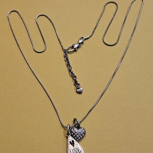 Puede incluir: Collar plateado con dos colgantes. Uno tiene forma de l&aacute;grima con la palabra "LOVE" y un dise&ntilde;o de coraz&oacute;n. El otro es un peque&ntilde;o coraz&oacute;n cubierto de cristales. El collar est&aacute; sobre un fondo dorado.