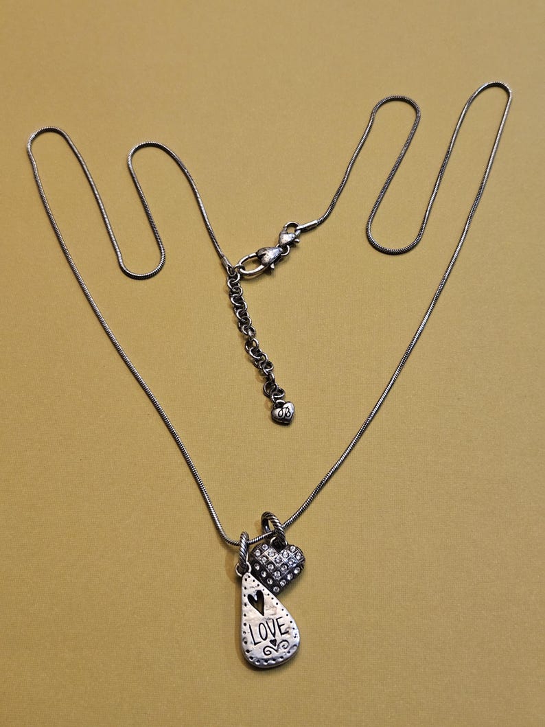 Puede incluir: Collar plateado con dos colgantes. Uno tiene forma de l&aacute;grima con la palabra "LOVE" y un dise&ntilde;o de coraz&oacute;n. El otro es un peque&ntilde;o coraz&oacute;n cubierto de cristales. El collar est&aacute; sobre un fondo dorado.